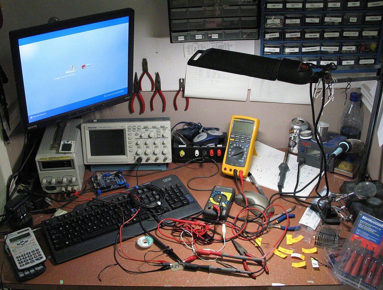 A cluttered home workbench with a computer monitor, soldering iron, multimeter and hand tools